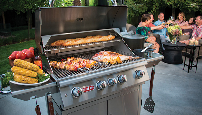 Family gathering around Bull BBQ grill with food cooking in an outdoor kitchen